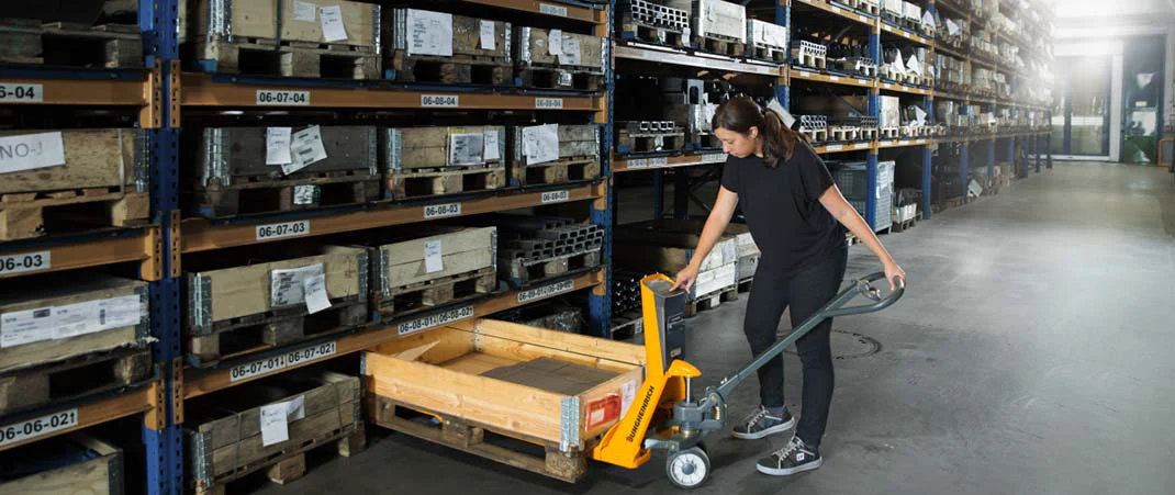 Frau schiebt einen Hubwagen unter eine Palette, um Waren aus einem Regal zu holen